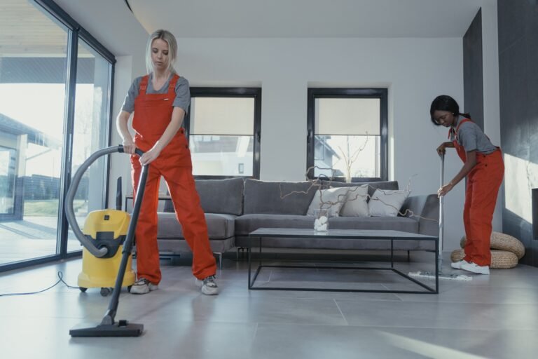 Two women in orange jumpsuits cleaning a modern home with a vacuum and mop.